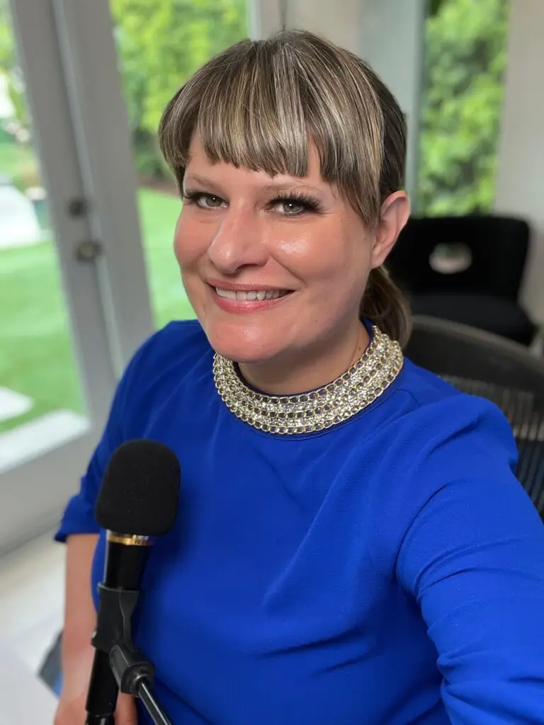 Charissa Nielsen smiling in a recording studio, wearing a royal blue top and gold collar necklace, speaking into a handheld microphone by a window.